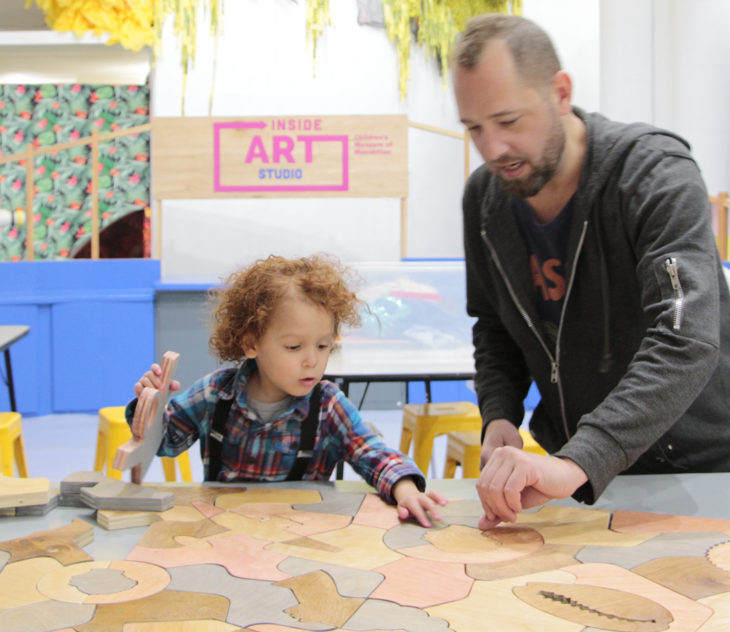 A child and his father interact with a table-sized wooden puzzle created by artist Damien Davis. Some pieces of the puzzle are shaped like coins with profiles on them, a woman in a hair wrap, a cowry shell, and a hoodie. 